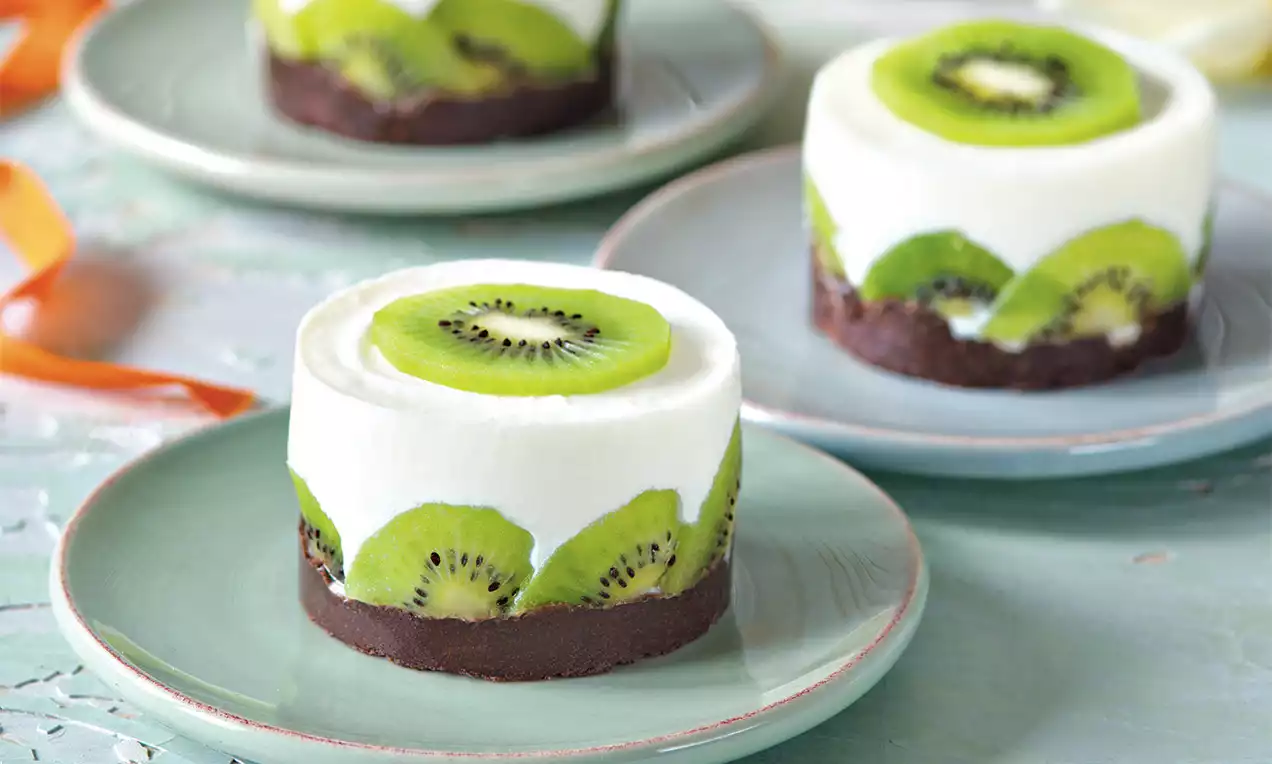 Mini tortičky na jemne zelenom tanieri, s bielou krémovou vrstvou, čerstvými plátkami kiwi a čokoládovým základom, usporiadané na svetlom povrchu