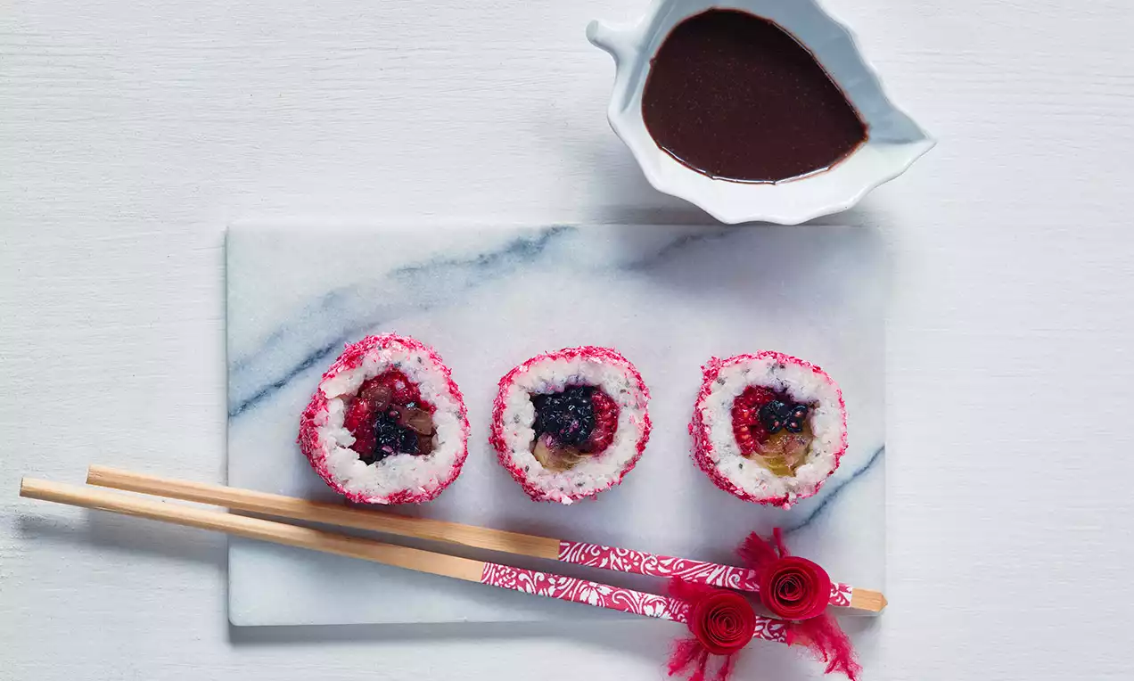 Tri farebné rolky sladkého sushi s kokosovým obalom a ovocnou náplňou, podávané na mramorovej doske s paličkami a čokoládovou omáčkou