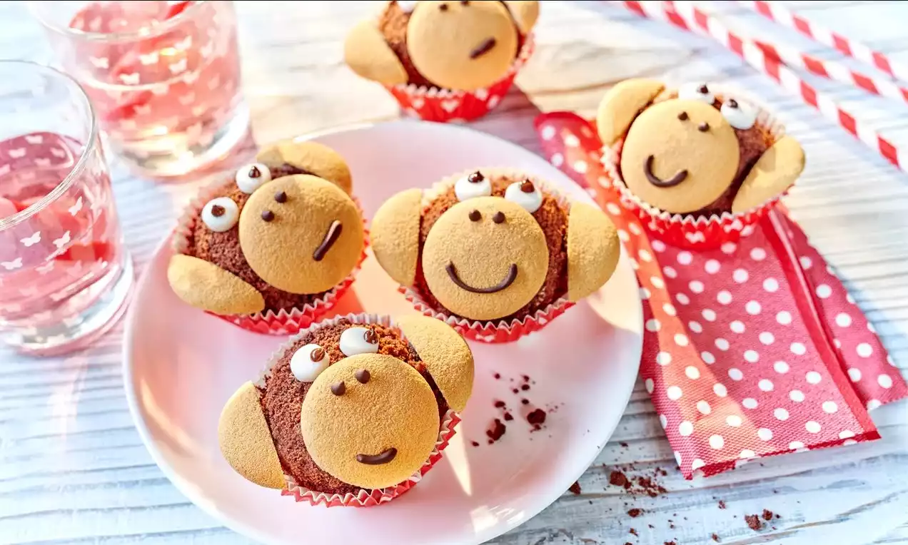 Čokoládové muffiny s jemne drobivým povrchom, ozdobené sušienkami v tvare opičích tvárí v červených bodkovaných košíčkoch na bielom tanieri