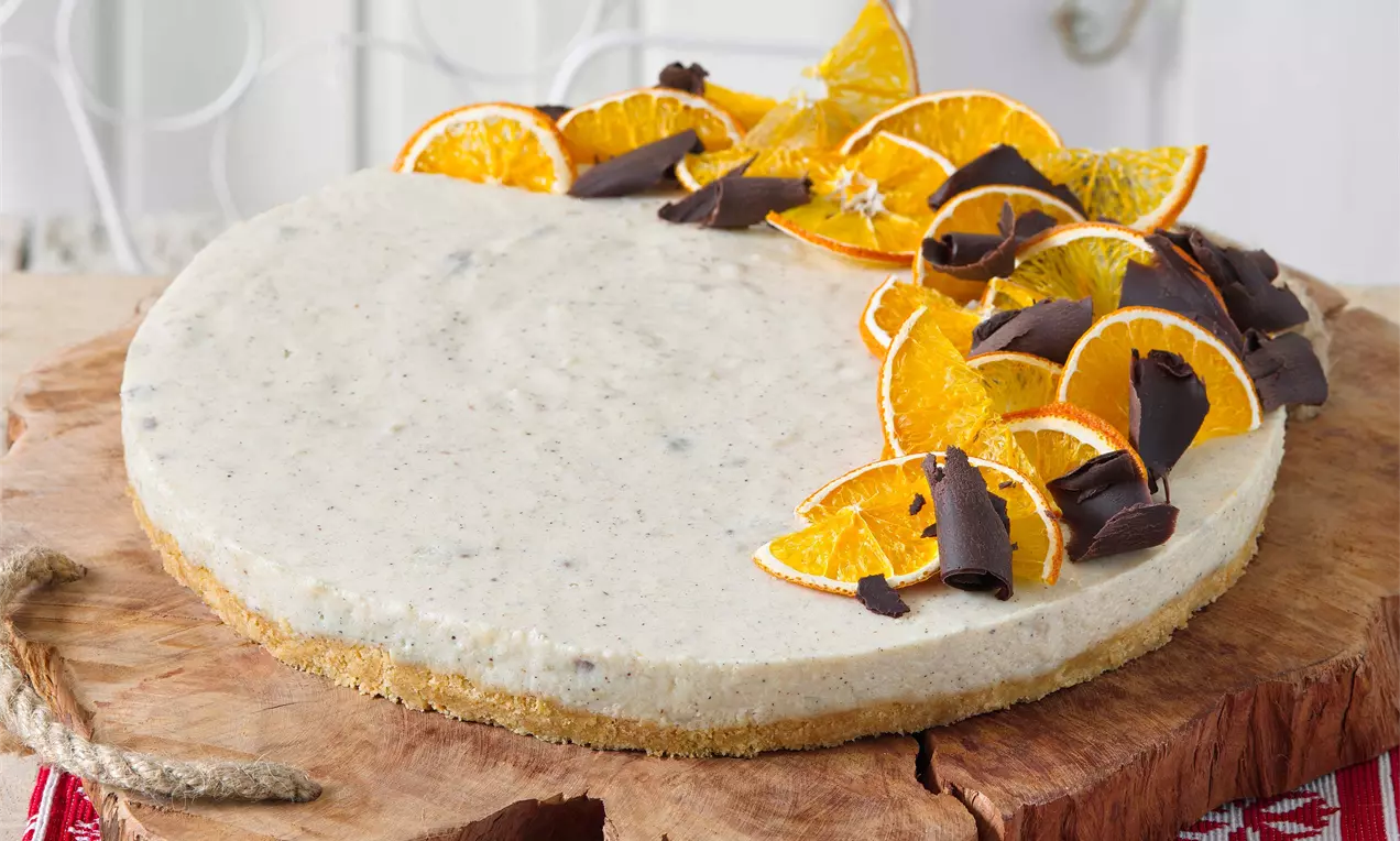Svetlý krémový cheesecake s jemnou textúrou na zlatohnedom základe, ozdobený plátkami oranžových pomarančov a kúskami tmavej čokolády