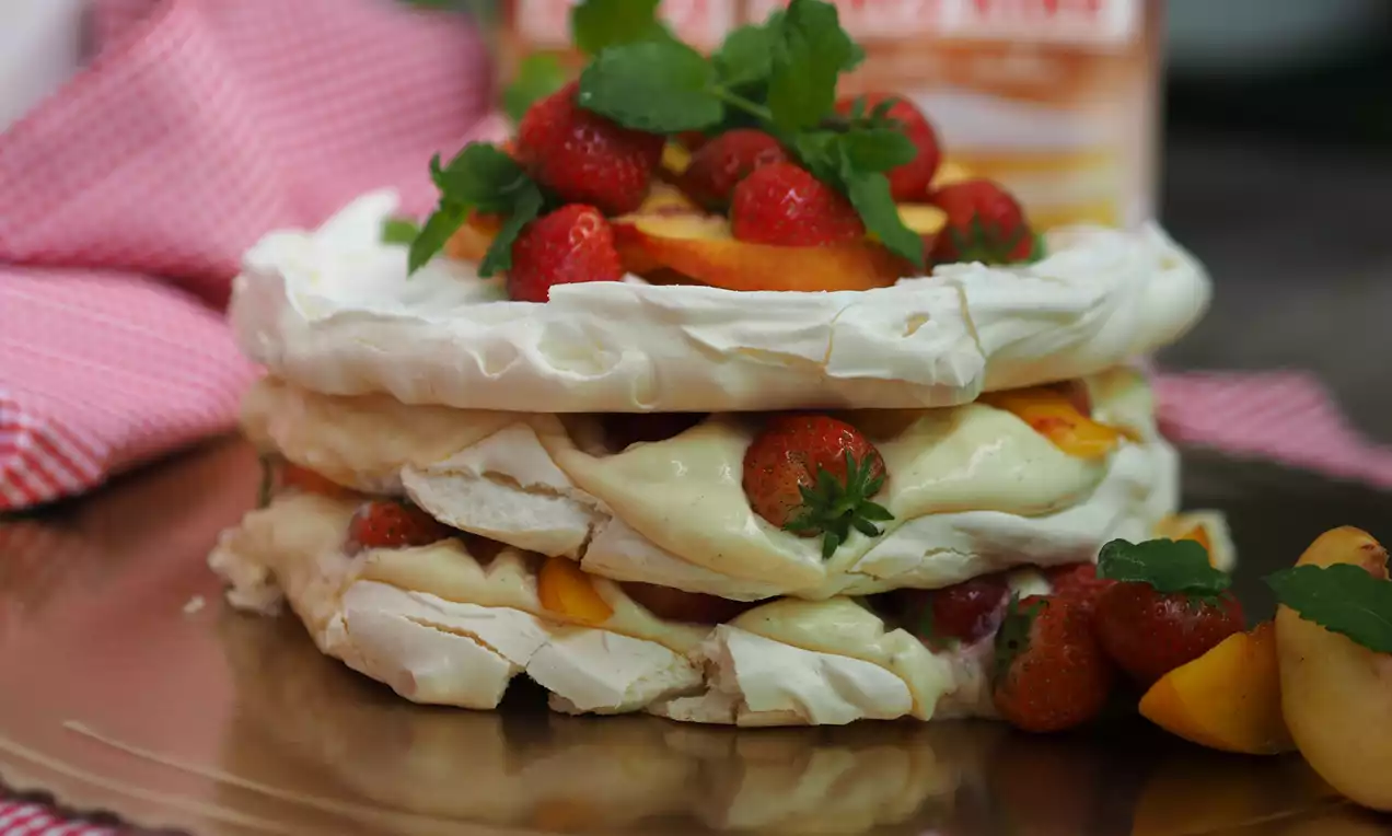 Dvojvrstvová biela pavlova s krémovo žltou plnkou, zdobená červenými jahodami, plátkami marhúľ a zelenými lístkami na zlatom podnose