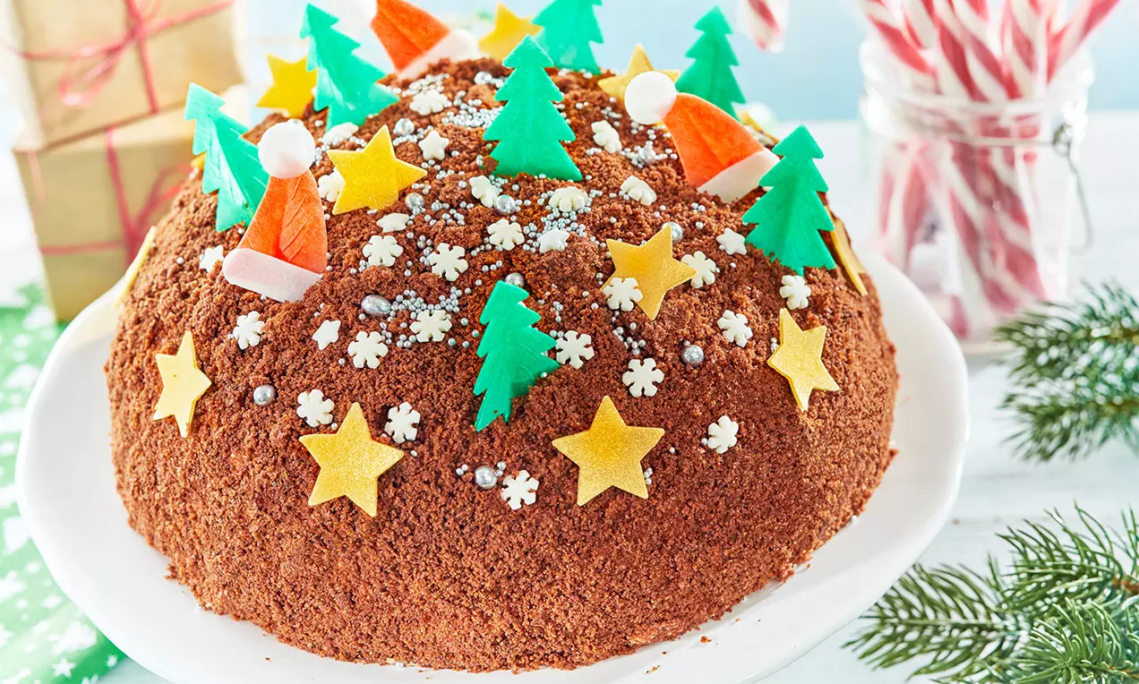 Kopulovitá torta z tmavočokoládových omrviniek, ozdobená farebnými hviezdami, stromčekmi a figúrkami na bielom tanieri so zimnou výzdobou