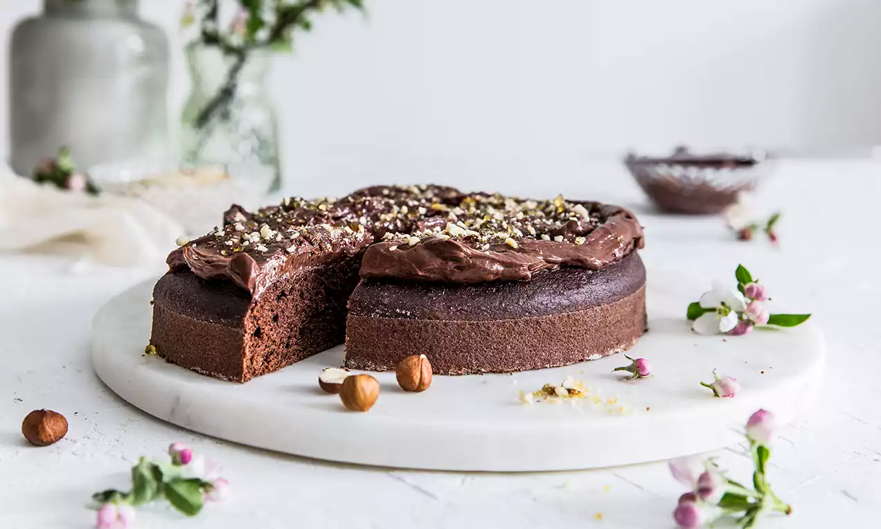 Vegan čokoládová torta tmavohnedého odtieňa s jemným krémom na vrchu, posypaná sekanými orechmi, položená na bielom mramorovom podnose medzi kvetmi a lieskovcami