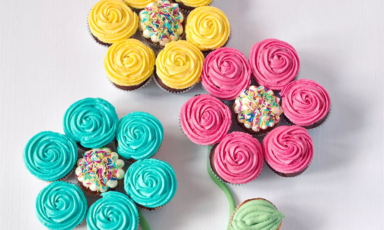 Muffiny s farebným krémom usporiadané do tvaru kvetov, žlté, ružové a modré okvetné lístky, stredy zdobené cukrovými posýpkami, zelené stopky