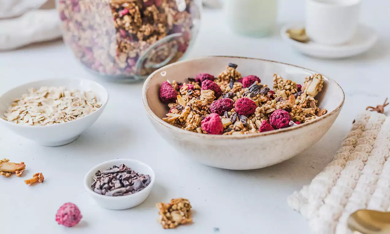 Granola zlatohnedá s textúrou drobných zhlukov, posypaná rubínovo červenými malinami v krémovo bielej keramickej miske na svetlom pozadí