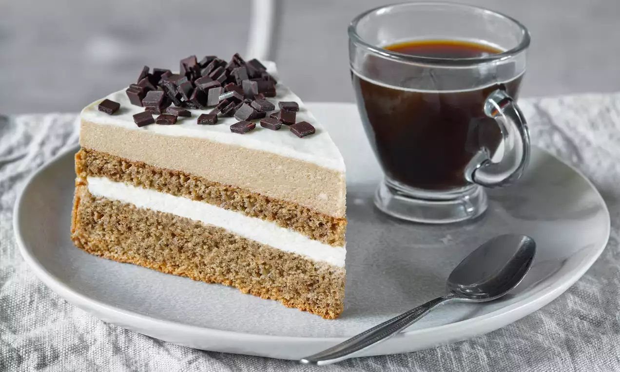 Cappuccino torta s vrstveným krémom a nadýchaným cestom, posypaná kúskami tmavej čokolády na šedom tanieri vedľa šálky čiernej kávy