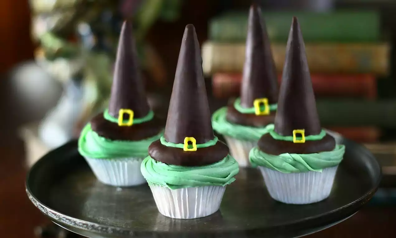Tmavohnedé čokoládové klobúky s malými žltými prackami na krémových zelenkavých cupcakeoch v strieborných košíčkoch na tmavom podnose