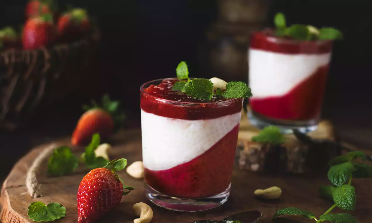 Dvojvrstvový dezert v pohári s rubínovo červenou jahodovou omáčkou, svetlými tapioca perlami, lístkami mäty a celými kešu na drevenom podnose