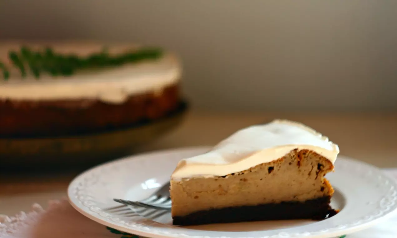 Plátok gaštanového cheesecaku s hladkou béžovou plnkou a tmavohnedým základom na bielom tanieri s jemným kvetinovým okrajom.