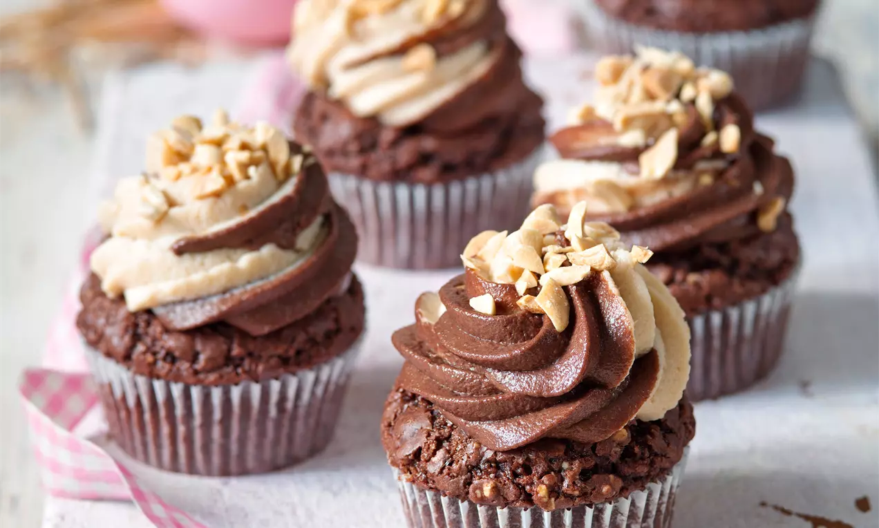 Čokoládové cupcakes s hlbokohnedým korpusom, špirálou krémovej polevy v odtieňoch hnedkastej, posypané nasekanými arašidmi na bielom podklade