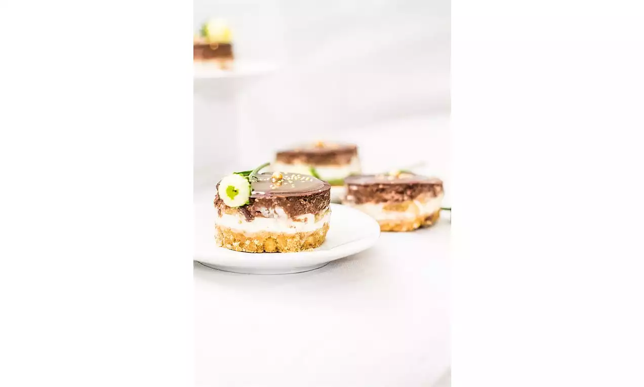 Dvojfarebný cheesecake s krémovou bielou vrstvou pod hnedým čokoládovým povrchom na sušienkovom základe, ozdobený kvetom na bielom tanieri