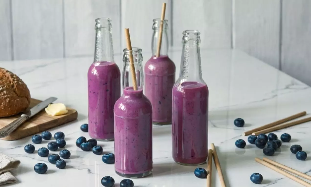 Proteínové smoothie sýtej fialovej farby v sklenených fľašiach so slamkami, obklopené čučoriedkami na bielom mramorovom stole, AI-labeled image