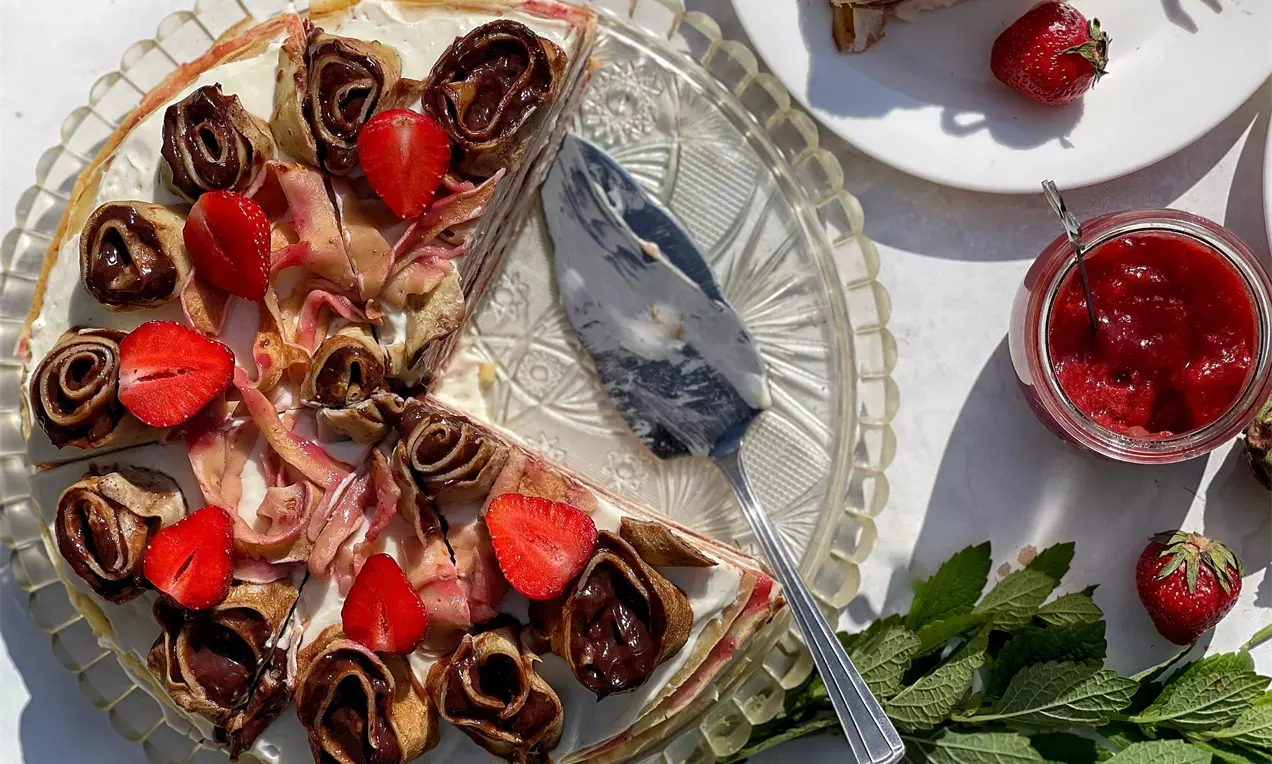 Palacinková torta s krémovou plnkou, vrstvami svetlého cesta, dekorovaná zrolovanými kakaovými palacinkami a čerstvými jahodami na sklenenom tanieri