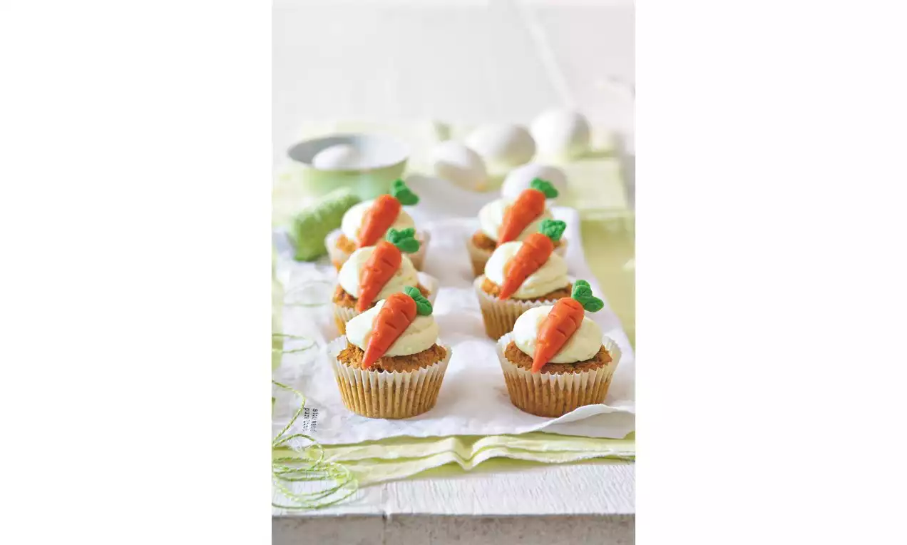 Mrkvové muffiny v zlatistých košíčkoch, zdobené hladkým bielym krémom a oranžovými cukrovými mrkvami so zelenými lístkami na bielej servítke