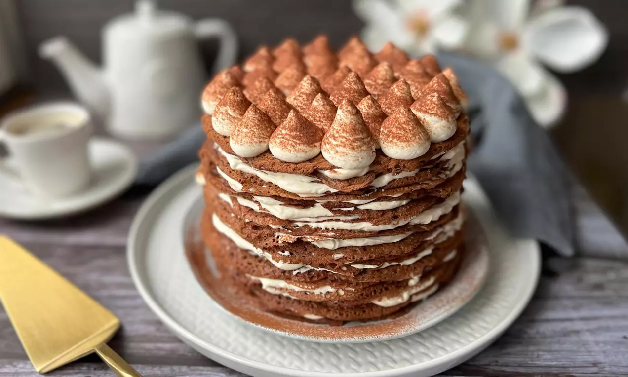 Vrstvená palacinková torta tmavohnedej farby s bledou mascarpone plnkou a vrstvou kakaom posypaných kopčekov na bielom tanieri