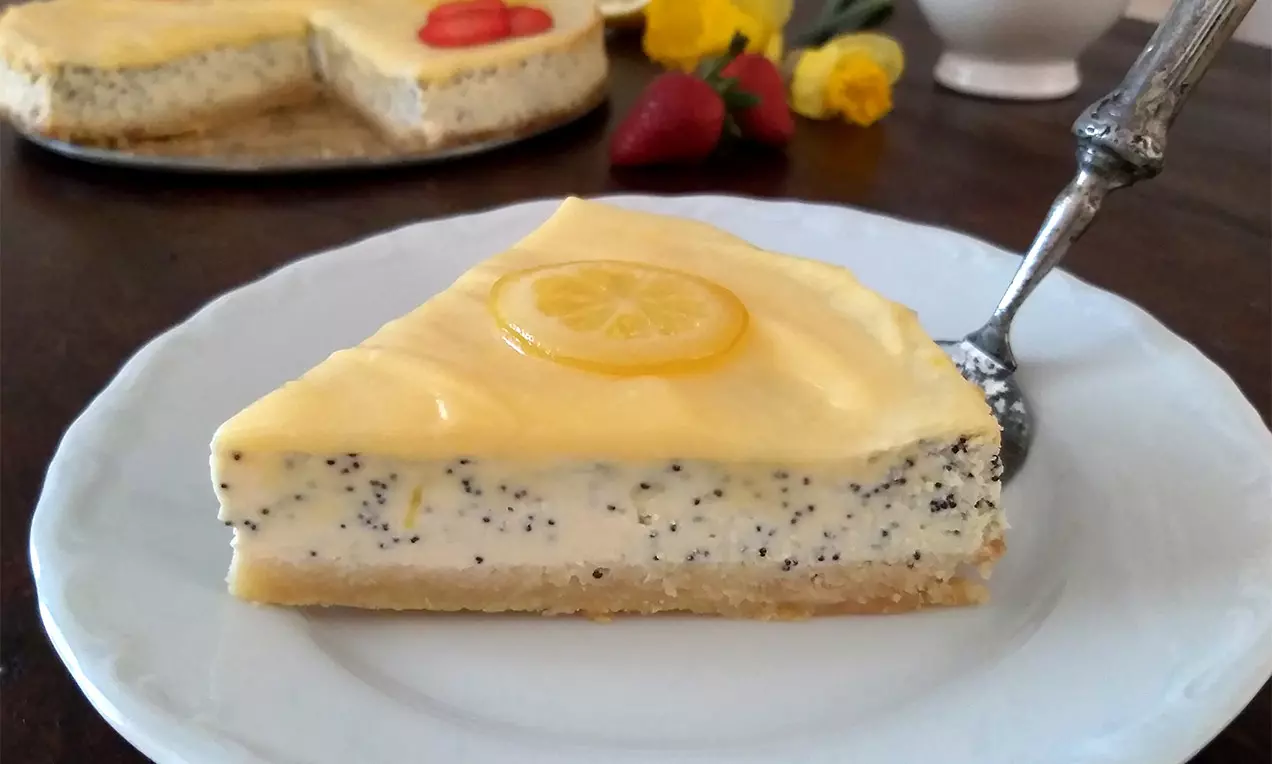 Makový cheesecake s hladkou svetložltou polevou, zdobený plátkom citróna, na jemnom sušienkovom základe podávaný na bielom tanieri
