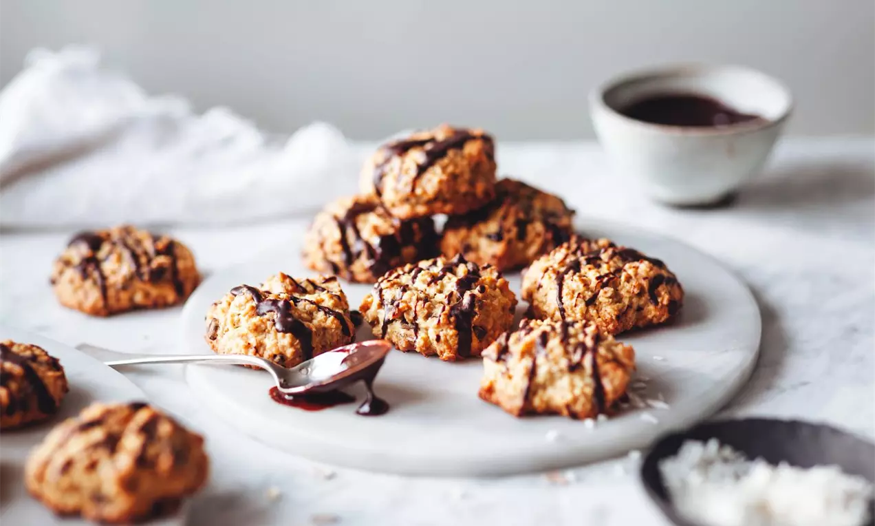 Chrumkavé ovsené cookies v nepravidelných tvaroch zlatohnedej farby, poliate čokoládou, usporiadané na mramorovej doske so šálkou čokolády.